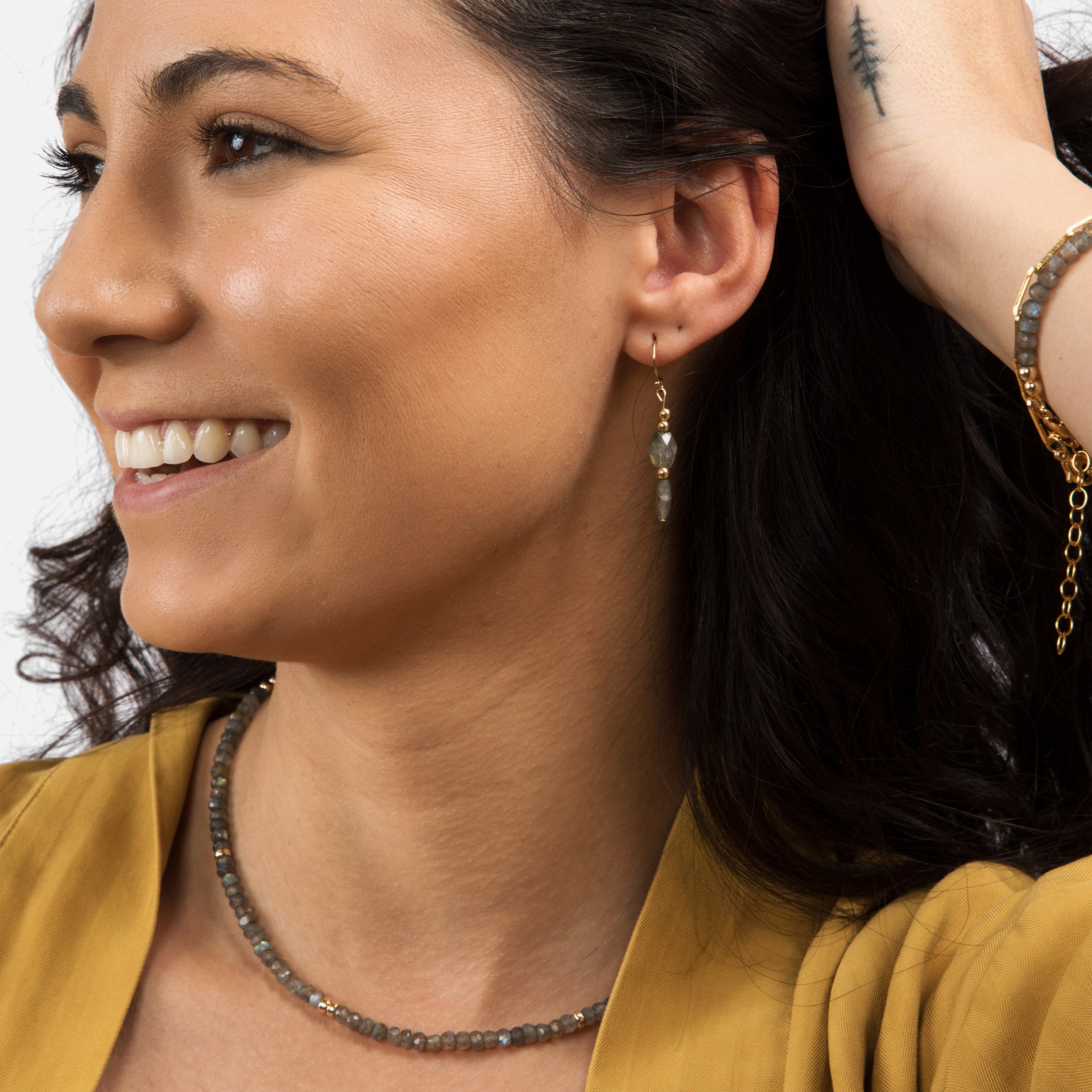 Model smiling with hands holding her hair, featuring stunning shimmering blue labradorite earrings, a matching necklace, and bracelet, creating a sophisticated and timeless look.