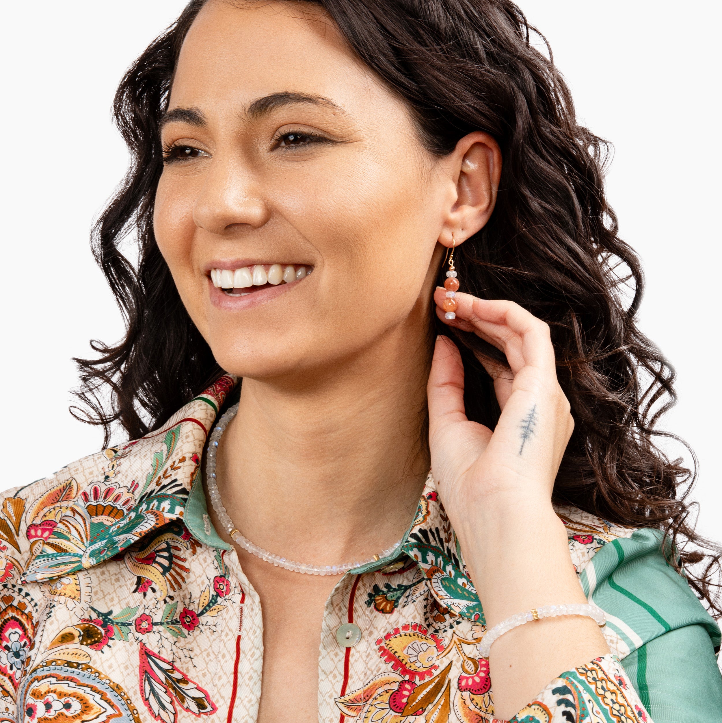 Model smiling with hand showing off Redondo earrings, necklace, and bracelet, along with a close-up of a stunning high-flash moonstone bracelet, adding the perfect touch of elegance from day to night.