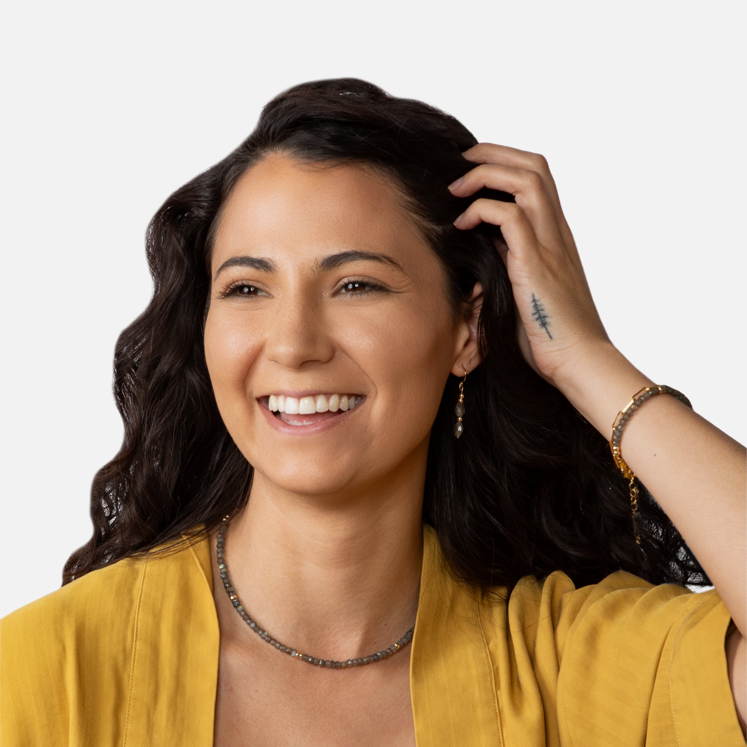 A cheerful model with long dark hair smiles and poses with the Serenity Collection. She is wearing tunnel faceted-shaped earrings, a delicate faceted rondelle bracelet, and a matching necklace, all featuring 14K gold-filled components that are tarnish-resistant. The jewelry’s unique texture and lustrous shine are highlighted against her elegant attire and natural makeup.