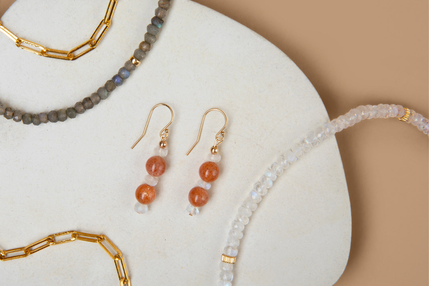 Flat lay of jewelry featuring a labradorite beaded necklace, a gold paperclip chain bracelet, a pair of earrings with sunstone and moonstone beads, and a moonstone beaded necklace, arranged on a white stone slab against a beige background.