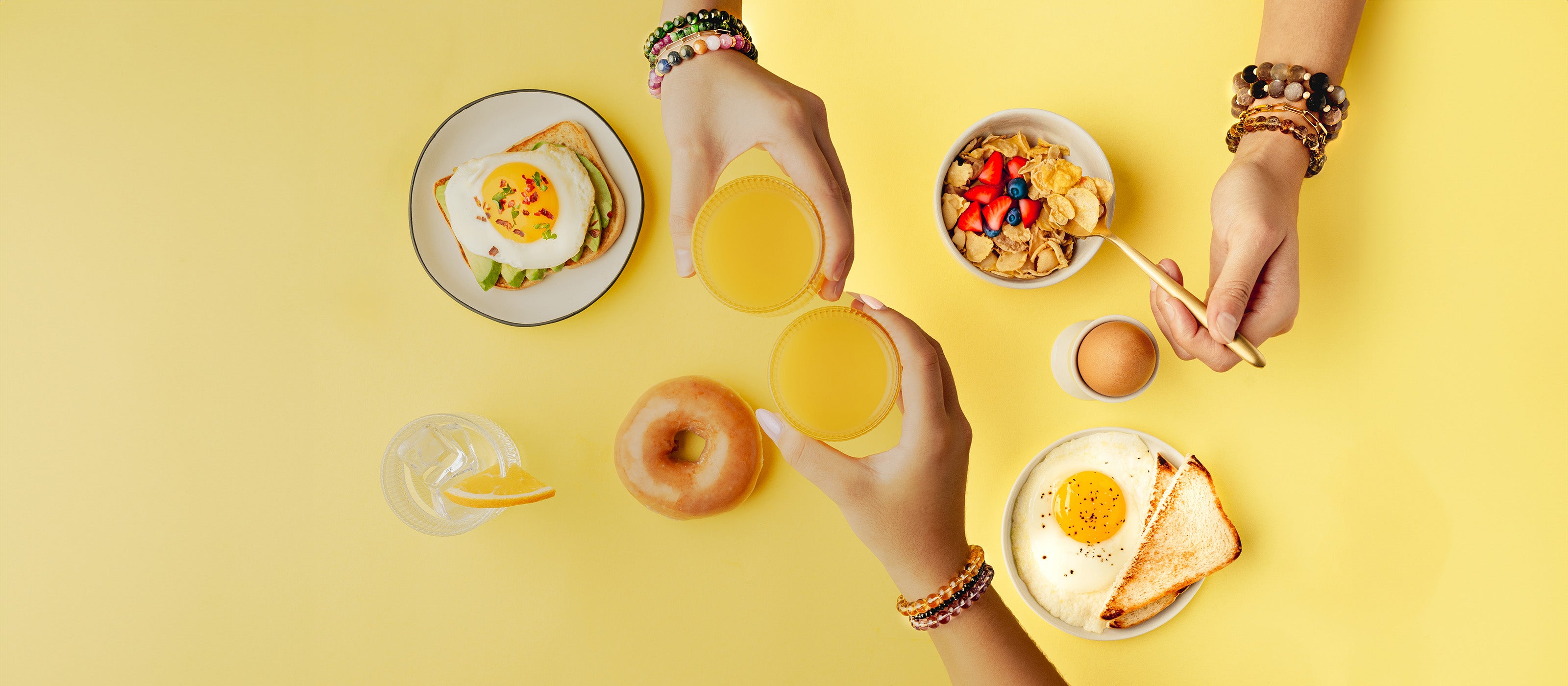 Overhead view of a breakfast scene with two people toasting with orange juice. Both wear colorful stacked beaded bracelets. The table features avocado toast with an egg, a bowl of cereal with berries, a donut, a glass of water with orange, and a plate with a fried egg and toast.