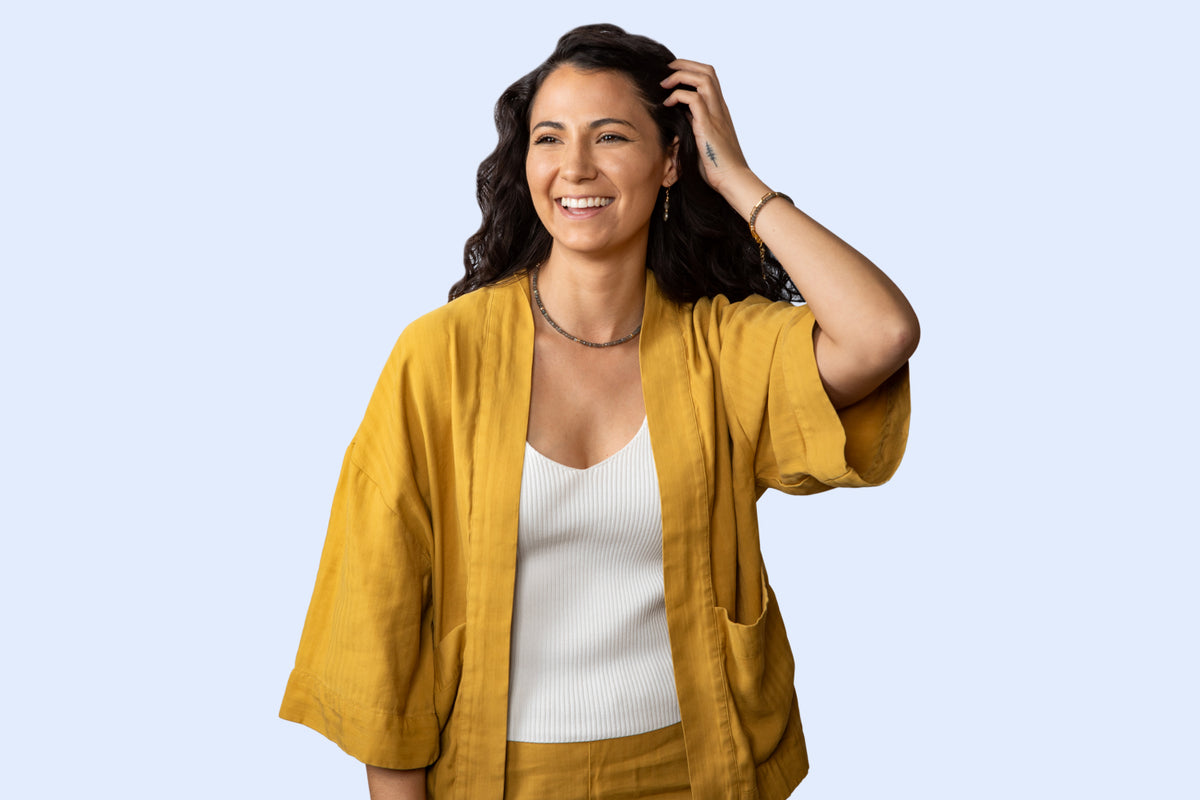 A cheerful model with long dark hair smiles and poses with the Serenity Collection. She is wearing tunnel faceted-shaped earrings, a delicate faceted rondelle bracelet, and a matching necklace, all featuring 14K gold-filled components that are tarnish-resistant. The jewelry’s unique texture and lustrous shine are highlighted against her elegant attire and natural makeup. Light blue background.