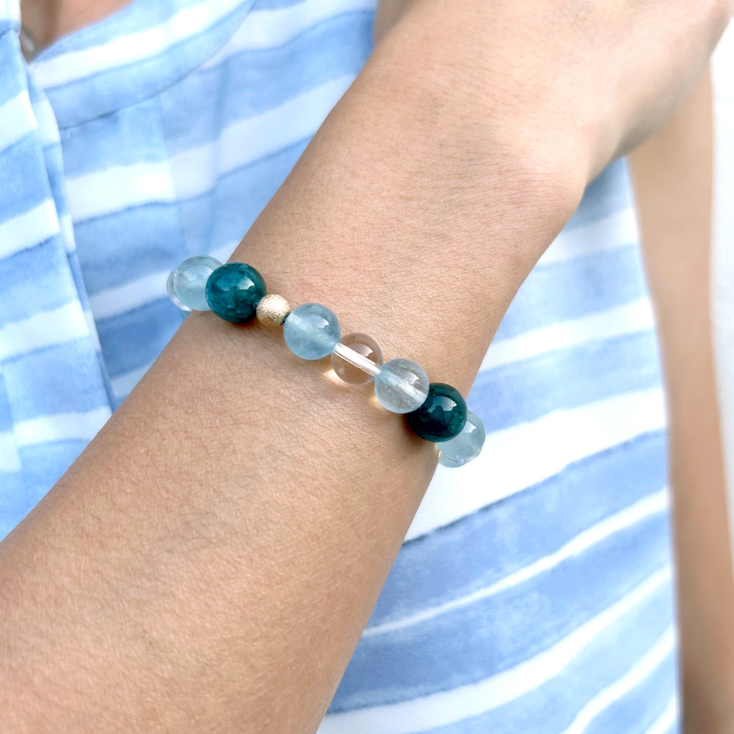 Close-up of a model's wrist wearing a bracelet with electric blue apatite, clear blue aquamarine, and clear quartz beads, set against a light blue stripe shirt, showcasing the ocean-inspired beauty of the piece.