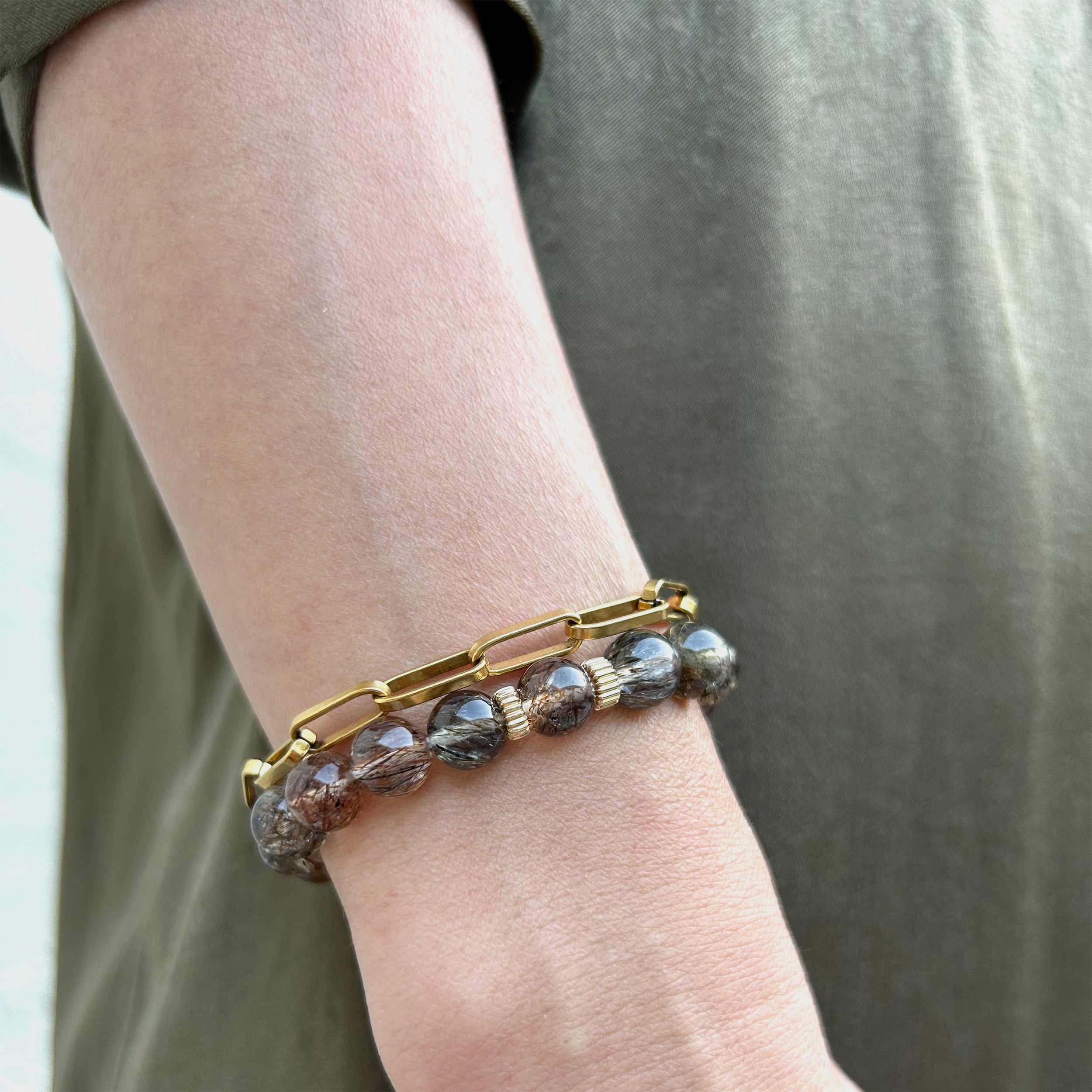 Close-up of a model wearing two bracelets: one with a gold paperclip design and another showcasing quartz with intricate patterns of tiny black copper-colored rutile needles, creating a classic and elegant style, highlighting its versatility