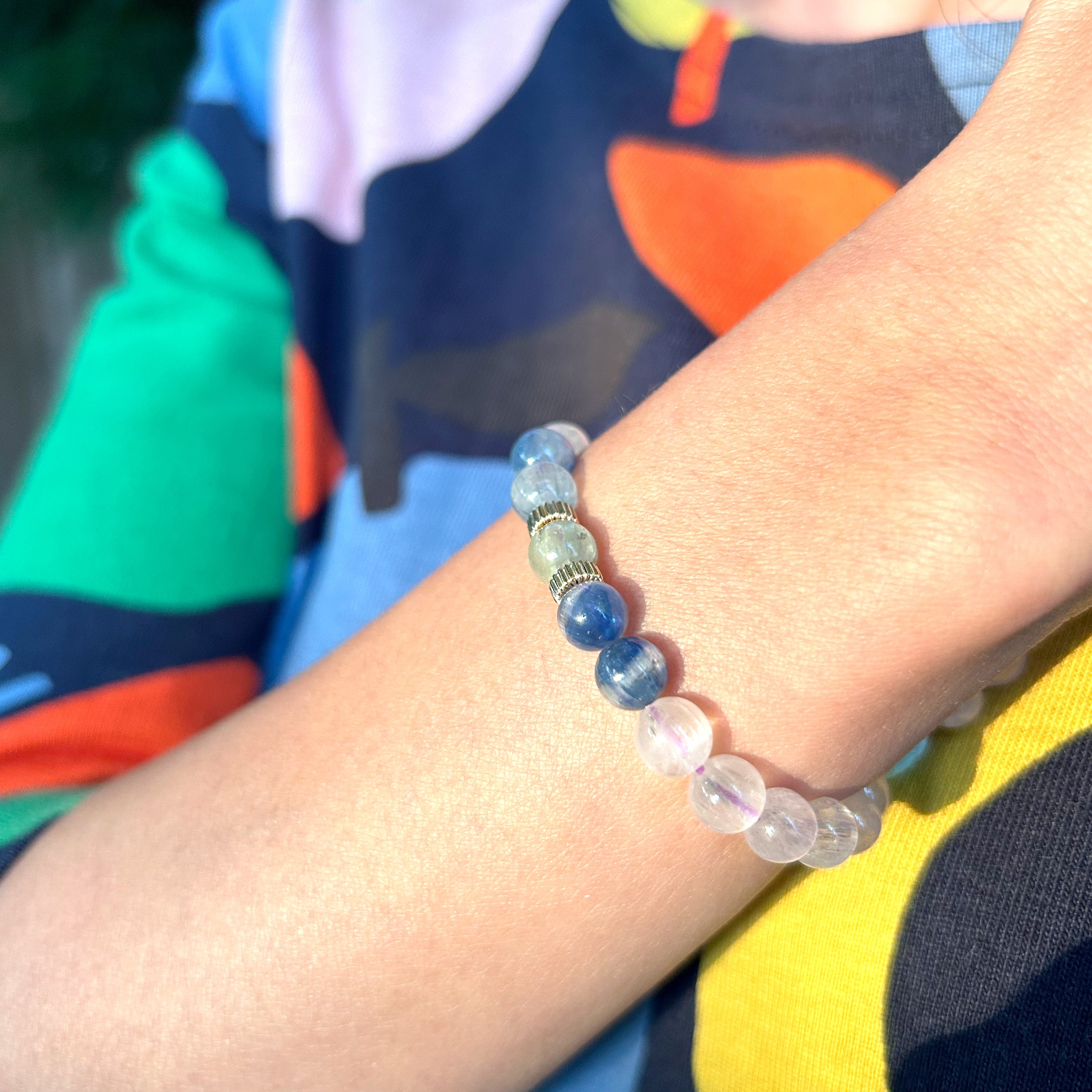 Close-up of a model's wrist wearing a bracelet showcasing rare cat eye kunzite gemstones with natural exterior lines, complemented by blue and green kyanite, creating a lustrous and elegant look against a colorful dress.