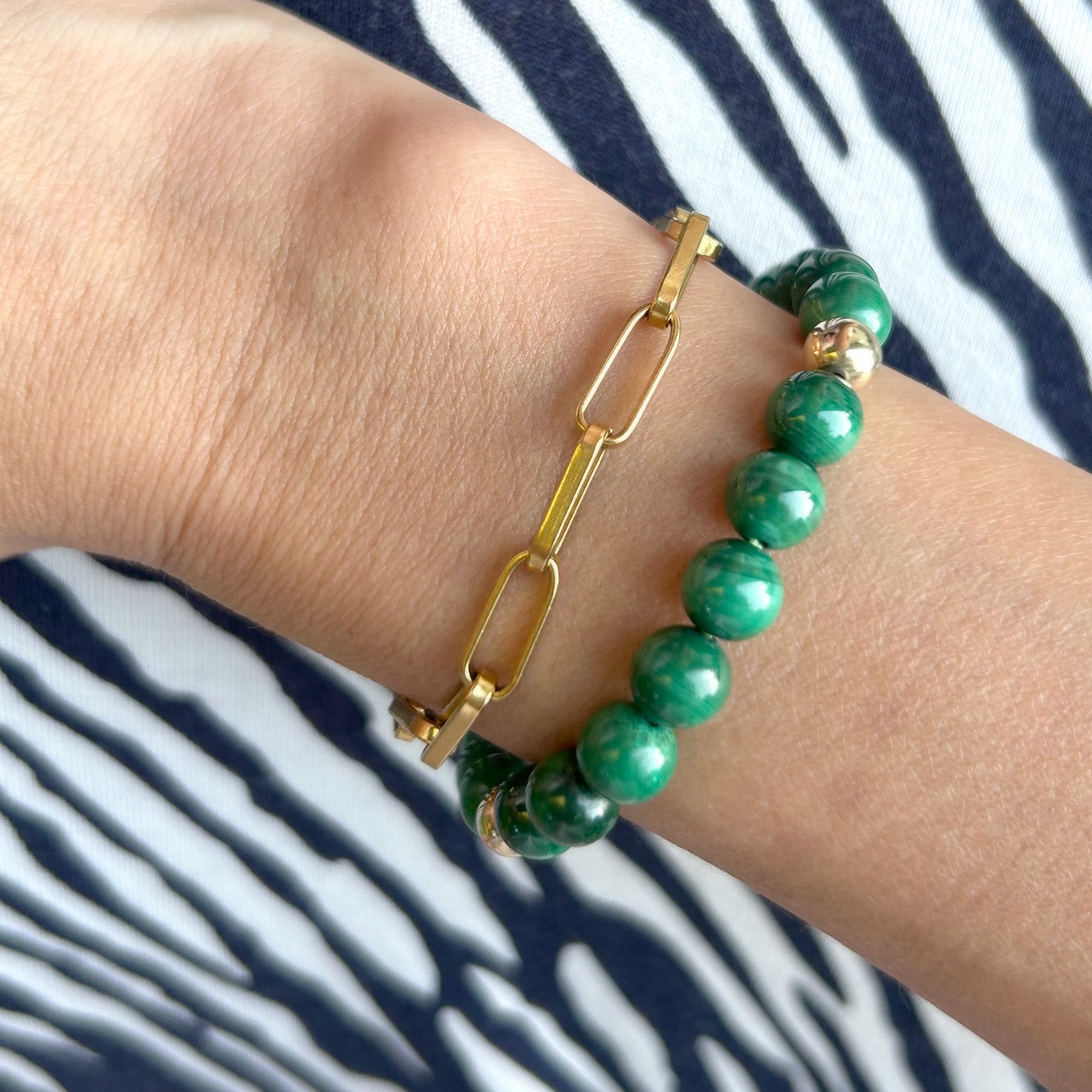 Close-up of a model's wrist wearing a rich green malachite bracelet, highlighting its beautiful banding and plume-like patterns, paired with other gold jewelry.