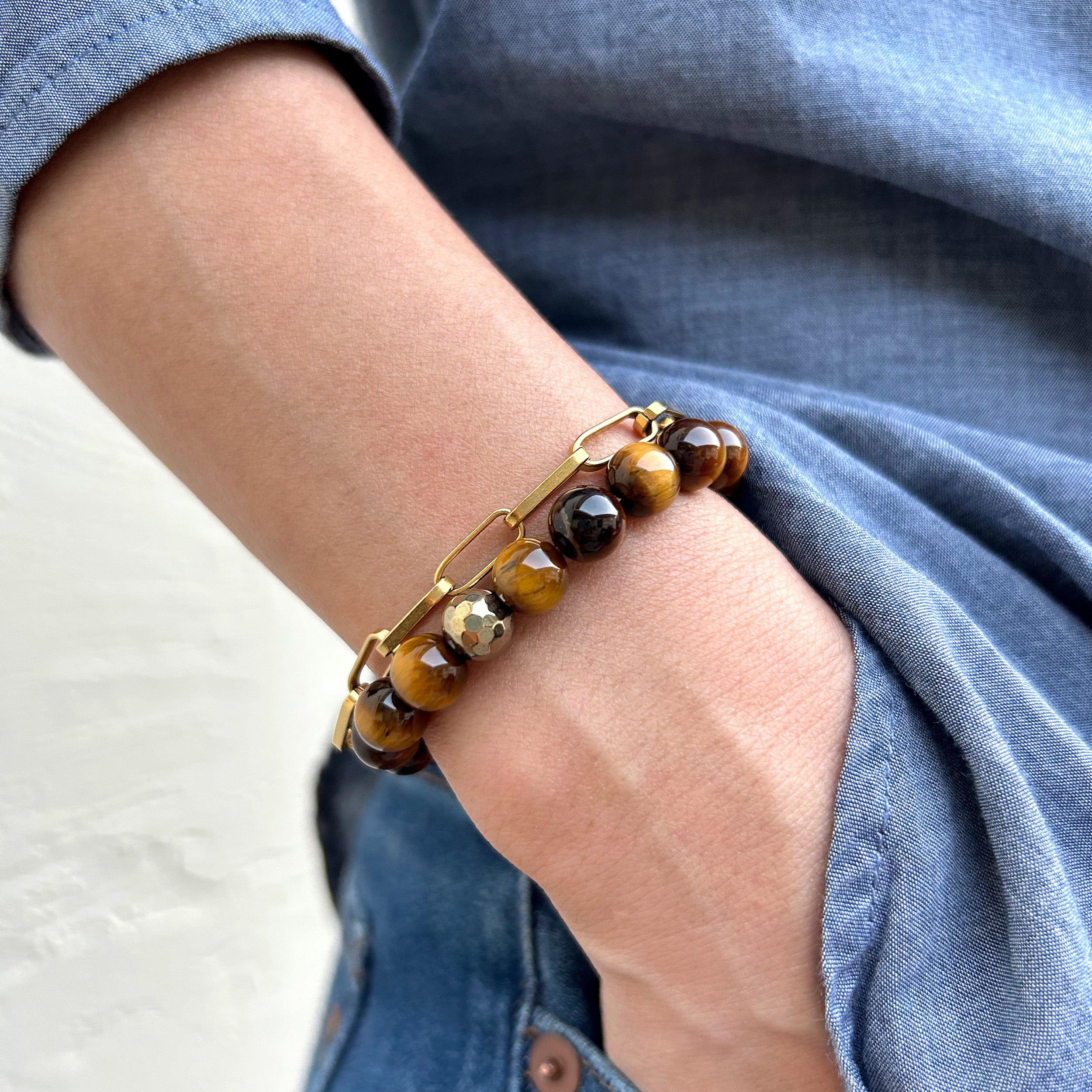 Close-up of a model's wrist wearing the bracelet, hands in pockets, highlighting the reddish-brown tiger eye, black obsidian beads, and metallic pyrite for a powerful and resilient look.